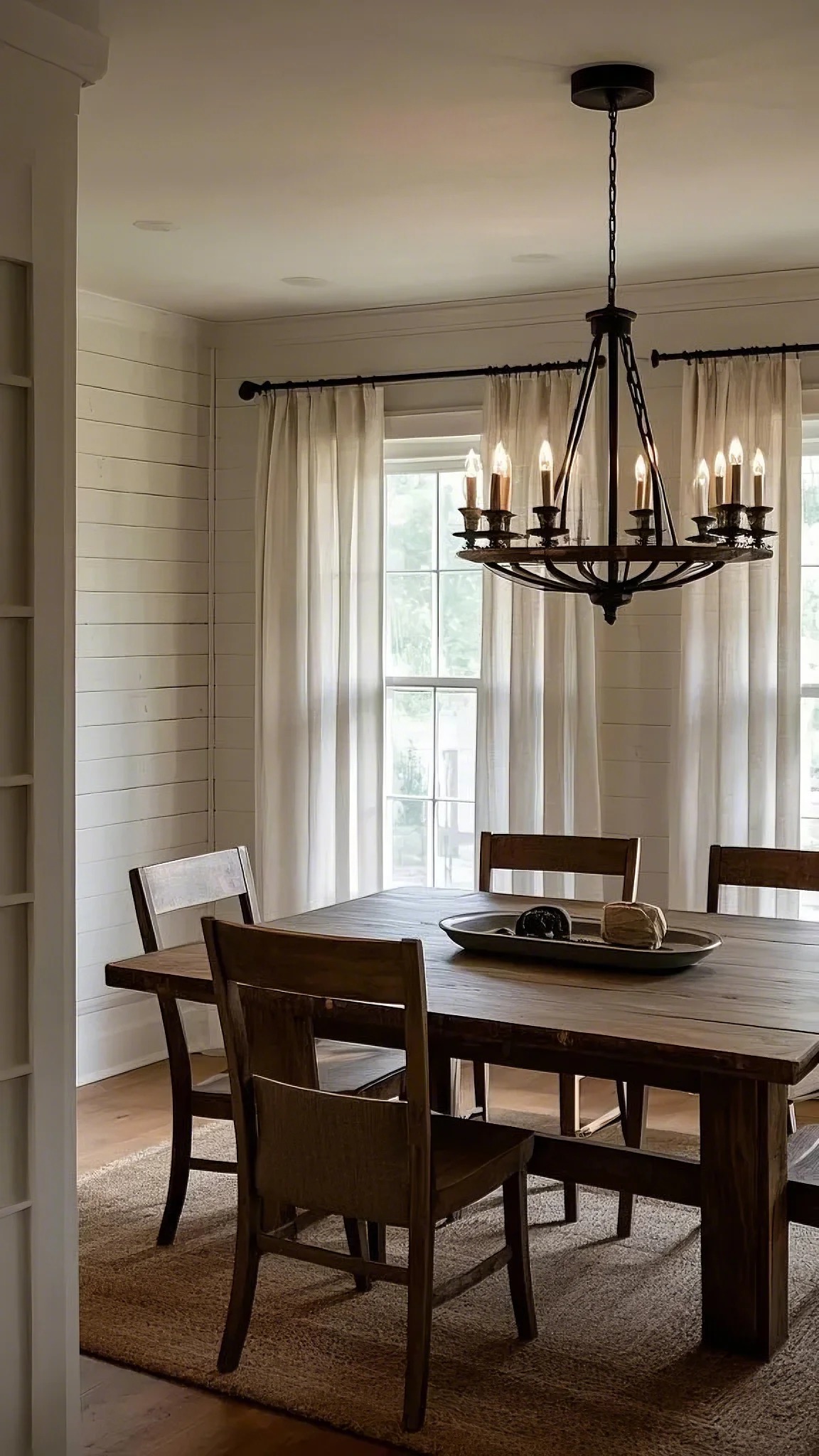 Cozy dining room with a rustic wooden table, four chairs, and a large iron chandelier. Soft sunlight filters through sheer curtains, creating a warm, inviting atmosphere.